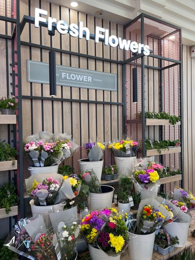 The image depicts a well-lit flower display area, possibly a florist shop or a section within a larger store. A prominent white neon sign above reads "Fresh Flowers". Below the sign, a grey plaque states "The FLOWER Gallery". The display features numerous bouquets of various colorful flowers, including pink, yellow, purple, and white varieties, presented in white buckets and pots. Some plants are also visible on tiered shelving units in the background. The backdrop consists of vertical wooden slats and a dark metal frame structure with wire mesh. The scene is clean and organized, suggesting a commercial setting focused on floral arrangements and plants. The provided crops offer closer views of the signage and different flower arrangements. There are no people visible in the image. The city of Donegal, Ireland is not visually indicated in the image.