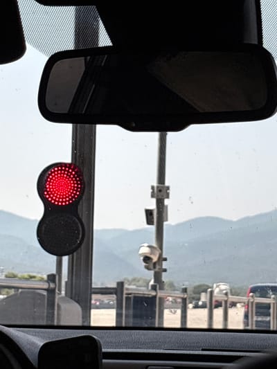 The image, taken from inside a vehicle in Le Boulou, France, shows a view through the windshield. A traffic light, displaying a red light, is prominently featured on the left side of the frame. The traffic light is mounted on a dark vertical pole, which extends upwards out of view. Behind the traffic light and pole, a set of distant, hazy mountains can be seen against a bright sky. A rear-view mirror is visible at the top of the windshield, reflecting a dimly lit interior. Lower down, a security camera is mounted on the same pole as the traffic light. In the background, a blurred scene of other vehicles and fencing suggests a road or parking area.