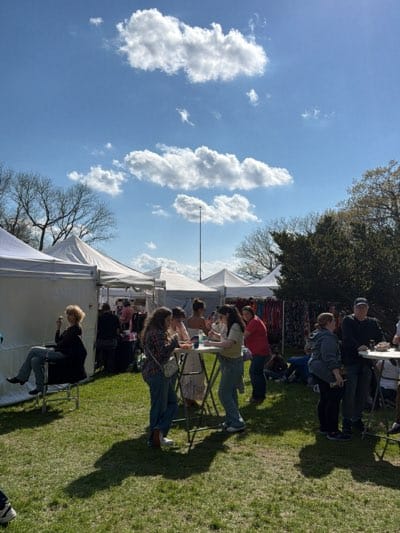 This image captures a vibrant outdoor market or festival taking place on a sunny day. The scene is set on a lush green lawn, likely a park or open field, with numerous white canopy tents scattered throughout the background, suggesting vendors or activity stations.
The weather is clear and bright, with a vivid blue sky dotted with scattered white cumulus clouds. Direct sunlight casts distinct shadows of people and objects on the grass, indicating a daytime event, likely mid-morning or afternoon. Bare-branched trees are visible in the distance to the left, while denser, green foliage can be seen to the right, suggesting a transition season like spring or early autumn in Rochester, United States. A tall, thin pole stands vertically in the mid-background against the sky.
Several people are present, engaging in various activities. To the far left, a woman with short blonde hair sits in a black folding chair, appearing relaxed as she holds something to her mouth. In the center-midground, a group of casually dressed young adults stands around a round, high-top table, engrossed in conversation, some holding cups. Another group of people, including a man in a black cap, occupies a similar table towards the right of the frame. Other attendees can be seen milling about among the tents in the background, some standing and others appearing to be seated on the ground.
The overall atmosphere appears social and relaxed, with people enjoying the outdoor setting and event. No visible text or specific branding is discernible within the image.