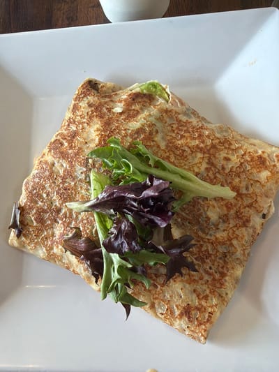 A close-up, overhead shot captures a savory crepe presented on a white square plate. The crepe is folded in half and filled with what appears to be avocado and shredded cheese, peeking out from the opening. A small garnish of mixed greens, including light green lettuce and darker red lettuce, sits atop the crepe. The crepe itself is golden brown with a slightly crispy texture, showing small bubbles from the cooking process. The white plate is clean, with a subtle shadow cast from the crepe, suggesting a light source from the top right. The background is blurred, showing a dark wooden surface on the left, possibly a table. The scene is indoors, likely a restaurant or home setting, with natural light illuminating the food. There is no visible text in the image.