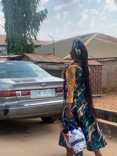 The scene depicts a street in Egbedi, Nasarawa State, Nigeria, under a partly cloudy daytime sky. In the foreground, a person, viewed from behind and slightly to the left, walks or stands on an unpaved, reddish-brown dirt surface. The individual has dark skin, wears a long, patterned dress featuring a teal, brown, and gold design with a golden lace long sleeve visible on the right arm, and a matching or complementary headwrap. Long, dark braided hair extends down the back, with some strands showing reddish tips. A dark-colored wristwatch is present on the left wrist. The individual carries two bags in the left hand: a white bag with a blue floral or abstract pattern and a smaller, translucent red bucket-style bag.
To the left of the person is the rear-left section of a silver/light gray Toyota Camry sedan. The car displays a Nigerian license plate: "NASARAWA AWE-349LB". The rear bumper shows significant peeling paint, revealing dark underlying material, and the left taillight area has a faded or damaged pinkish patch.
In the mid-ground and background, several buildings with corrugated metal roofs, mostly brown and rusty, are visible. One building on the left has a partially obscured sign that reads "VET GA..." or "VET GK..." and "COMMUNICATION". A tall tree with dense green foliage stands to the far left, partially obscuring the upper portion of a building. A utility pole or street light standard with an upward-curving arm is visible between the buildings and the person.