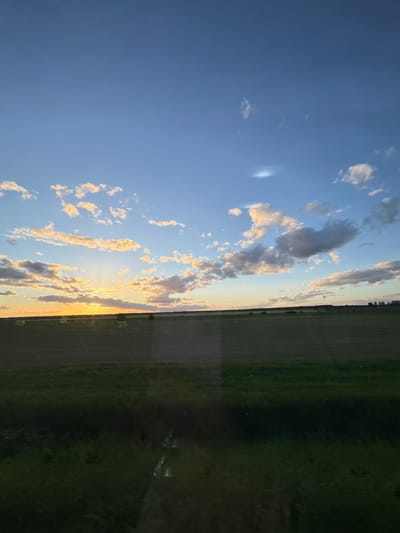 A photograph depicts a landscape at the Мажэйкаўскі сельскі Савет. The left side of the image features a blue sky with scattered clouds illuminated by the sun. The right side shows a green field.