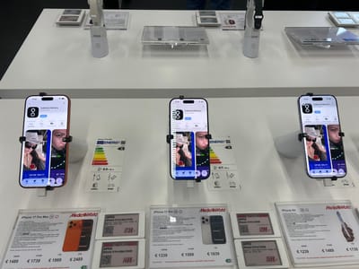 The image depicts a well-lit, white retail display counter within a MediaWorld store located in Lipomo, Italy. Three smartphones, identified as iPhones (specifically, iPhone 17 Pro Max, iPhone 17 Pro, and iPhone Air models based on adjacent price cards), are showcased vertically, secured by black, two-pronged security mounts.
Each phone's screen displays the identical interface of an app store page for "Collective Memory," a social network application with a 5.0-star rating and a 16+ age recommendation. The app's interface shows two distinct image previews: one featuring a person in a white bucket hat with "JACQUEMUS" text and sunglasses, and another of a person making a pouting or "duck face" expression.
Beneath the phones, the white counter holds various clear acrylic product information cards and digital price tags. These cards detail specifications, storage capacities (256GB, 512GB, 1TB, 2TB), and prices in Euros for the displayed iPhone models, with prices ranging from €1239 to €2489. Several cards also feature "Energia" labels indicating energy efficiency (Class A) and estimated battery life (e.g., 53h 0min, 47h 0min), alongside symbols for features like 1000x zoom and IP68 water resistance. Digital tags prominently display "APPLE" and "TOP SELLER" alongside prices.
In the upper background, additional white security stands and information cards are visible, suggesting other products such as smartwatches are also on display. The overall environment is a clean, organized electronics retail setting.