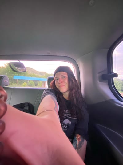 A young person with long, dark, wavy hair and a black beanie is sitting in the backseat of a vehicle, taking a selfie. They are wearing a black t-shirt with a skull graphic and the words "DEAD INSIDE" visible. The person has tattoos on their arms. The interior of the vehicle is visible, including the gray leather seats, a rearview mirror reflecting a hilly landscape with a blue-roofed structure, and the vehicle's ceiling and side panels. The background outside the windows shows a slightly hazy, overcast sky and green foliage. The location is the city of Verkhneozernoye, Russia.