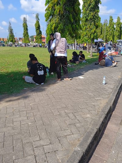 People are sitting on grass beside a paved area with water bottle. The background shows trees, vehicles, and buildings. The location is in Kebondalem, Indonesia.
