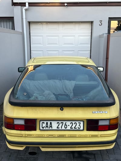 The image shows the rear of a yellow Porsche 944 parked in front of a house in Blouberg, South Africa. The house has a white garage door and the number "3" is visible above the door. The Porsche has a South African license plate that reads "CAA 276-223". The environment appears to be a residential street. There are no visible people in the image.