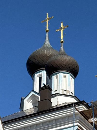 The image shows the upper portion of two onion domes on a building, likely a church, against a clear blue sky. Both domes are dark, with a textured surface, and topped with golden crosses. The dome on the left is slightly taller and positioned to the left of the dome on the right. The building below the domes is white with light blue trim and features rectangular window-like openings. A dark chimney is visible between the domes. Part of the roof and scaffolding are also present in the lower right of the image. The location is identified as St. Ozerki, Russia.