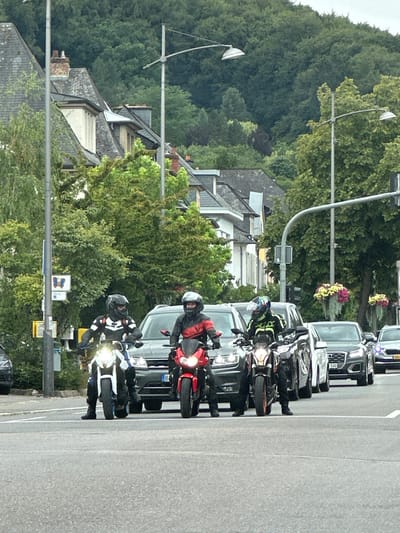 This is a daytime, eye-level, medium shot captured on a street in Echternach, Luxembourg. The image shows four motorcyclists on their bikes, stopped at what appears to be a traffic light or intersection. The motorcyclists are wearing full protective gear, including helmets. The first motorcyclist on the left is on a white motorcycle. The second motorcyclist, slightly to the right, is on a red motorcycle. The third motorcyclist, further right, is on a black and orange motorcycle. The fourth motorcyclist is on a black motorcycle. Several cars are visible behind and to the side of the motorcycles, including a dark grey Audi. The background features buildings with pitched roofs and trees, with a densely forested hillside in the distance. Hanging flower baskets are visible on the right side of the frame. The street surface is asphalt. The overall scene depicts a moment of pause in traffic on a town street.