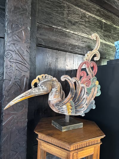 An intricately carved wooden bird statue is centrally positioned on an octagonal wooden display table within a structure in Tibubiyu, Indonesia. The statue, depicting a stylized bird with a long beak and ornate, scrolled tail feathers, exhibits weathered paint in shades of cream, brown, gold, red, dark grey, and teal. It is mounted on a dark rectangular base atop a slender dark rod. To the left, a substantial dark wooden pillar with deep, traditional Indonesian relief carvings featuring swirling patterns rises vertically. The background consists of dark wooden wall paneling, displaying visible grain and texture, some planks oriented horizontally and others vertically. A partial glimpse of a blue or teal object is visible on a dark surface in the upper right background. No people or specific actions are depicted.