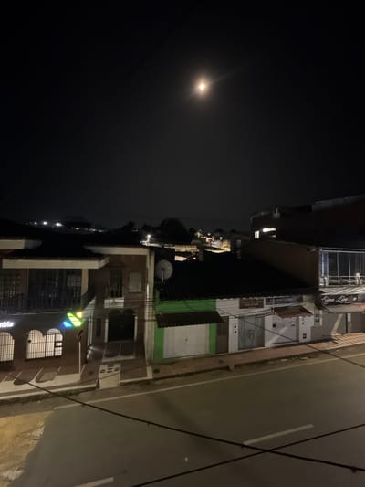 An elevated, nighttime perspective captures a street scene in Timaná, Colombia, illuminated by streetlights and a prominent full moon positioned in the upper center-right of the dark sky. The foreground features dark power lines crisscrossing the frame. Below these lines, a dark paved road with white lane markings runs horizontally across the image, appearing devoid of vehicles or pedestrians. A light-colored sidewalk separates the street from a row of commercial and residential buildings.

From left to right, the visible structures include:
1.  A two-story building with a light-colored facade, dark roof, and a ground-floor commercial space featuring gated windows and a partial sign displaying "ombia" and a multi-colored triangular logo (green, blue, yellow). The second story has a balcony with a dark railing.
2.  Adjacent to it, a single-story building with a green facade and a white roll-down security shutter. A dark, tiled roof with a mounted satellite dish is visible above this structure.
3.  To its right, a single-story white building with a dark grey security shutter. A sign above the shutter reads "TIENDA DE ROPA ONDAR." A connected section to its right, also white, has a similar dark grey shutter and a partial sign reading "CELEBRY CORNER."
4.  At the far right, a multi-story building features large reflective glass windows on an upper floor, and a sign on the lower facade reads "Lima Lima." The ground floor entrance is obscured by closed security shutters.

In the mid-ground and background, additional buildings and illuminated distant hills are visible as dark silhouettes against the night sky, indicating further urban or rural development. No individuals or active interactions are observed within the scene.