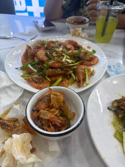This image captures a close-up view of a table laden with food and drinks, suggesting a meal is in progress. The primary subjects are plates of cooked shrimp, some whole and garnished with green onions, and a smaller bowl containing what appears to be discarded shrimp shells and some whole shrimp. There's also a white plate with some cooked food, possibly octopus or another seafood dish, with green onions. A clear plastic cup filled with a yellow-green beverage and a black straw is visible, held by a person whose hands and fingernails are partially in frame. The setting appears to be indoors, likely a restaurant, with a white tablecloth and some background elements that hint at a busy environment, including a blue and white graphic sign. The lighting suggests it could be daytime or evening. The overall mood seems casual and perhaps focused on the enjoyment of the meal, with the abundance of food and drink. There is no discernible text in the image.