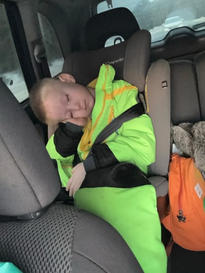 The image depicts the interior of a passenger vehicle in the city of Зюкайка, Russia. A young child, dressed in a neon green and black winter jacket, is observed asleep in a grey Capella-branded child safety seat in the rear. The child is secured by a black seatbelt and has their head leaning towards their right shoulder, resting against their arm. Adjacent to the child on the right are an orange backpack with a black zipper and a brown fuzzy object. Through the vehicle's rear window, an exterior scene is visible, showing a distant vehicle on what appears to be a road under an overcast or snowy sky.