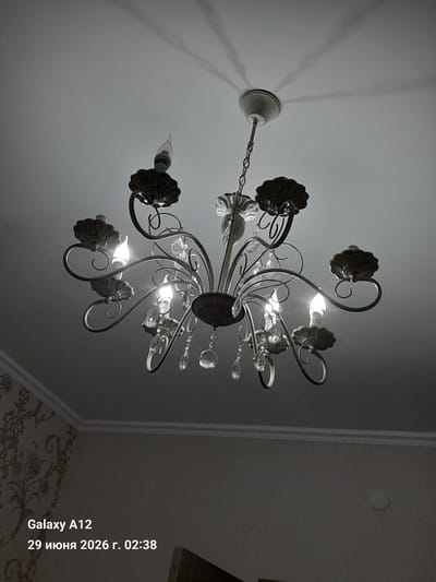 A dimly lit, black and white, low-angle interior shot captures a vintage-style chandelier hanging from a textured ceiling. The chandelier features ornate metalwork with multiple candle-shaped light bulbs, some of which are illuminated. Crystal teardrop-shaped pendants hang from the arms of the chandelier, reflecting the light. Shadows of the chandelier are cast on the ceiling. In the lower left corner, overlaid text reads "Galaxy A12" and "29 июня 2026 г. 02:38". The bottom portion of the image shows a section of a wall with patterned wallpaper and a decorative ceiling molding. The scene is devoid of people or any discernible activity.