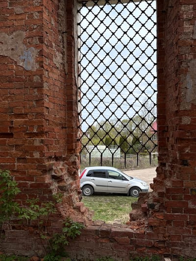 The image shows a weathered brick structure, likely a remnant of a building, with a decorative metal grille partially filling an opening. Through the opening, a white car is visible parked on a patch of grass. The location is Staroe Rakomo, Russia. The scene is still, with no visible people or actions. The environment appears overgrown and possibly neglected.