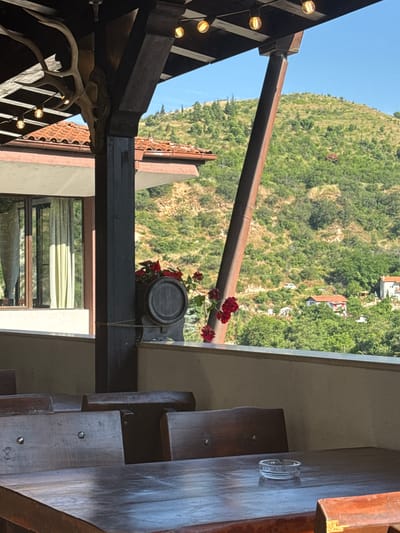 This image depicts an outdoor seating area, likely a restaurant or hotel terrace, located in Melnik, Bulgaria, during daytime under a clear blue sky. The immediate foreground features empty, dark-stained rustic wooden tables and chairs. On the table in the lower right, a clear glass ashtray is present. Overhead, a dark wooden beam structure, possibly part of a pergola or roof, is visible, from which string lights with exposed incandescent bulbs hang. Attached to one of the vertical wooden posts supporting this structure is a large set of animal antlers, including a partial skull. A tall, reddish-brown metallic or wooden support pillar angles upwards and to the right, also connecting to the overhead structure. Along the balcony railing, a small wooden barrel-shaped planter, filled with vibrant red flowers, is secured with twine. To the left, part of a building with a terracotta-tiled roof and a window with light-colored curtains is visible. The background showcases a panoramic view of a steep, verdant mountain covered in dense green trees and shrubs. Scattered within the hillside vegetation are several small houses with reddish-orange tiled roofs, and a narrow dirt road winds through the foliage, on which a white van is partially visible. No individuals are present in the scene.