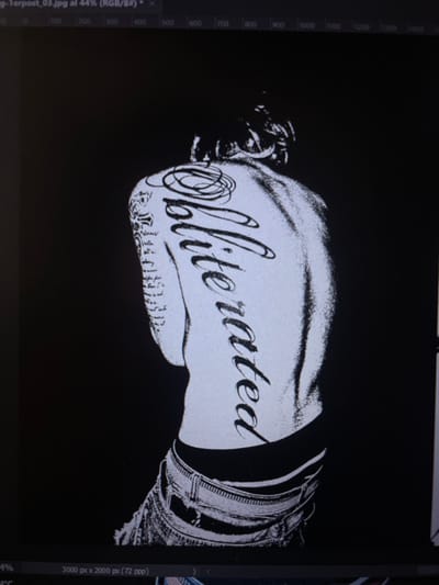 A black and white image features the word "Obliterated" in an elegant, cursive font, positioned over what appears to be a person's torso. The torso is adorned with tattoos, adding intricate detail to the design. The background is solid black, creating a stark contrast that emphasizes the central figure and the text. The image's style suggests it may be a design for a t-shirt or similar merchandise.