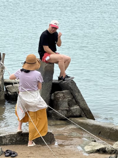 Two people relax by the water on a stone structure. A man in a black shirt, red shorts, sneakers, and a baseball cap lies back with legs crossed. A woman wearing a lilac shirt, a straw hat, and an orange skirt with a white layer stands closer to the water, leaning on a rope. Two pairs of sandals sit on the ground nearby. The water stretches out in the background.