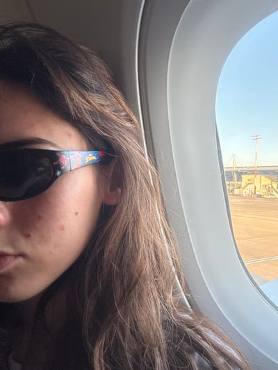 The image displays a close-up, angled view from inside an aircraft. On the left, a person's upper body is partially visible, primarily their left profile and shoulder-length brown hair. The individual is wearing dark sunglasses, the right temple of which features a colorful "Spider-Man" graphic. Minor skin blemishes are present on the visible portion of the person's face, specifically on the cheek and near the nose. To the right, an oval aircraft window frames an exterior scene under a clear blue sky. An airport tarmac, light brown in color, is visible, with various ground vehicles and structures, including what appears to be the wingtip of another aircraft in the lower right. In the distance, a large white cable-stayed bridge with distinctive A-frame pylons is discernible, along with a red and white striped pole on the tarmac. The setting is an airport within the Sdot Dan Regional Council, Israel.