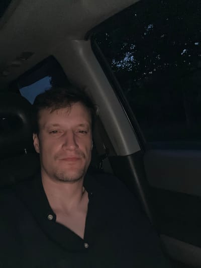 A man is lying down in the back seat of a car, with his head resting on the seat. He is looking at the camera with a slight smile. The scene is dark, suggesting it is nighttime or evening. Outside the car window, there appears to be water, possibly from rain, with reflections of lights. The man is wearing a dark colored shirt with buttons. The interior of the car is dimly lit, with some reflections visible on the surfaces. The image has a candid feel, as if taken by a passenger in the front seat. The location context provided is Makiv, Ukraine, but this is not visually evident from the image itself.
