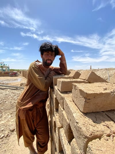 The image depicts a man in Deh-E Bagh, Afghanistan, leaning against a stack of mud bricks under a partly cloudy sky. He is wearing a traditional brown shalwar kameez and has a hand on his head while sticking his tongue out. The setting appears to be a brickyard, with a water-soaked ground and a motorcycle in the background, near a structure with flags. A tall, chimney-like structure is visible in the distance.