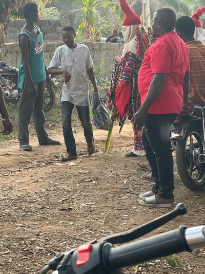 The scene depicts a daytime gathering on an unpaved dirt path in Umueze, Nigeria. Six individuals are visible, including one prominently dressed in an elaborate masquerade costume. In the foreground, the black handlebar and red kill switch of a motorcycle are partially visible, angled towards the left.
From left to right:
* A partially visible person's arm and torso are on the far left.
* A young male stands next to a parked motorcycle, wearing a teal sleeveless jersey with "UJET" visible on the chest and dark grey trousers, paired with blue flip-flops.
* A male in a light grey long-sleeved shirt with a buttoned placket and dark jeans is in the center, holding a patterned black and white bag in his left hand and several green palm fronds or similar leaves in his right hand. He is gesturing downwards with his right hand and wears brown flip-flops.
* A person stands in an ornate, multi-colored costume, predominantly red and white, featuring a tall, conical red headpiece. The costume is adorned with intricate patterns, multiple rows of hanging tassels, and fringes.
* A heavy-set male stands to the right of the costumed figure, wearing a red short-sleeved t-shirt and dark trousers. He is positioned next to a motorcycle, with part of its wheel and chrome fender visible. He wears grey and brown sandals.
* Behind the red-shirted man, another person is partially visible, appearing to wear a brown striped shirt, and another individual in a similar red conical headpiece is also partly obscured.
The background features dense green vegetation, including palm trees, and a low concrete wall. Additional figures are vaguely visible in the distant background, seated or gathered amidst the foliage. The ground is dry earth with scattered leaves and debris.