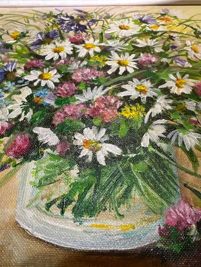 The image is a close-up, overhead shot of a painting depicting a vibrant bouquet of wildflowers in a white vase. The flowers are a mix of white daisies with yellow centers, small pink and purple flowers, and delicate blue blossoms, all surrounded by lush green foliage. The painting's texture, likely oil on canvas, is visible, with visible brushstrokes. The vase is simple and white, with a textured rim, and sits on a surface that appears to be a light brown tablecloth or tabletop. The background is a soft, muted beige. The painting is lit from above, casting subtle shadows. There are no people or animals present in the painting. The image does not provide any information that would allow for its location in Sergeevka, Russia.