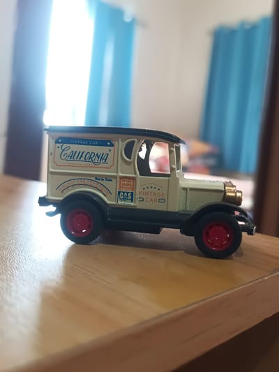 This is a close-up, eye-level shot of a vintage toy van. The van is primarily cream-colored with black trim around the roof and windows. It has red wheels with silver hubcaps. The side of the van features several colorful decals. The largest decal on the side reads "CALIFORNIA" in stylized blue lettering with an orange border, and below it, "RETRO CARS SHOWROOM" in smaller orange text. Another decal features a gas pump icon and the words "VINTAGE GAS STATION". The van is positioned on a light brown wooden surface, likely a table, with a slightly blurred background that includes blue curtains and bright natural light suggesting daytime. The van itself is the sole subject of the image, and there is no activity or event taking place. The visible text primarily relates to the "California" theme and "Vintage Cars." The overall impression is of a well-preserved collectible toy displayed indoors.