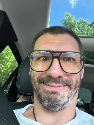 A man wearing large, square-framed glasses with a dark rim and yellow-tinted lenses takes a selfie inside a car. He has short, dark hair and a salt-and-pepper beard. His eyes are wide and looking towards the camera, and he is smiling broadly, showing his teeth. He is wearing a white t-shirt. In the background, through the car window and sunroof, lush green trees and a bright blue sky with a few wispy clouds are visible. The interior of the car is dimly lit, with the black leather headrest of a car seat visible to the left of the man. A child's hand can be seen reaching out from behind the headrest, towards the front of the car. The image appears to have been taken on a sunny day, likely during daylight hours. The exact location in Povedniki, Russia cannot be determined from the image.
