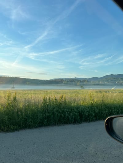 The image shows a landscape from a moving vehicle. The right side of the frame shows a gray asphalt road and the side mirror of the car. Adjacent to the road is a grassy area with tall green and yellow vegetation. Further into the landscape, there's a thick fog obscuring the view, but a treeline and a mountain range are visible in the background. The sky is blue with cirrus clouds. The location appears to be rural, possibly outside a city. There is no discernible evidence of Kastel, Bulgaria, within the frame. No people, specific subjects, actions, or interactions are discernible.