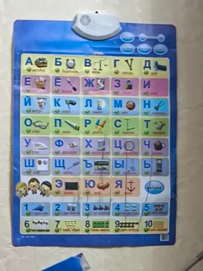 This is a vertical, eye-level photograph of a children's educational alphabet and number chart hanging on a light-colored, textured wall. The chart is predominantly blue with colorful sections for letters and numbers.

The chart displays the Russian alphabet, with each letter accompanied by an illustration and a word beginning with that letter. For example, the letter 'А' has an image of a bus, and the word "автобус" (avtobus - bus) below it. Other illustrations include a clock, a hammer, a glass, scissors, a teapot, and a phone. Below the alphabet section, there are rows of numbers from 1 to 10, each with corresponding illustrations. The numbers 1 through 5 feature images like an airplane, pencils, and dominoes. Numbers 6 through 10 have images of buttons, numbers of items (e.g., 7 buttons, 8 green blocks), and a blank rectangle for number 10.

At the top of the chart, there is a white electronic device with a speaker grille. Below the alphabet section, there is a row of cartoon drawings of children's faces. In the lower right corner, a button labeled "Музыка" (Muzyka - Music) is visible. A blue object, possibly a pen or marker, is partially visible in the bottom center of the frame.

The setting appears to be indoors, likely a home or classroom in Khasavyurt, Russia, given the context. The lighting suggests it is daytime, and the image is slightly blurry, indicating it was taken with a handheld device. The overall tone of the chart is cheerful and educational, aimed at young children.