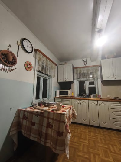 The image depicts the interior of a small, somewhat dated kitchen, likely in a rural setting within Навадворскі сельскі Савет, Belarus. The walls are painted in a split color scheme, with the lower half a pale blue and the upper half white. The ceiling is also white and appears to have exposed wooden beams, one of which has a bright light fixture attached, illuminating the space.
On the left wall, a circular analog clock with a black frame hangs above two decorative ceramic plates. The lower plate is larger and features a rustic, terracotta-colored pot, while the plate above it is smaller and adorned with a colorful, circular pattern.
A window with white wooden panes is situated in the center of the left wall. It is covered by a sheer, patterned curtain. The window overlooks what appears to be an outdoor, dark space.
In front of the window, a table is covered with a checkered tablecloth in shades of orange, brown, and white, featuring a repeating pattern of red fruit. On the table are several plates, bowls, a mug, and what looks like a bottle of nail polish.
The right side of the kitchen features white upper and lower cabinets. Above the window, a mounted deer head with antlers serves as a decorative piece. A microwave oven is placed on the countertop between the upper and lower cabinets. The countertop itself is a light wood color. On the countertop, near the window, are various mugs, cups, and what appears to be a kettle.
The floor is covered with a parquet flooring arranged in a geometric pattern of light brown and darker brown wooden tiles. The overall impression is one of a lived-in, functional kitchen, likely belonging to a private residence.