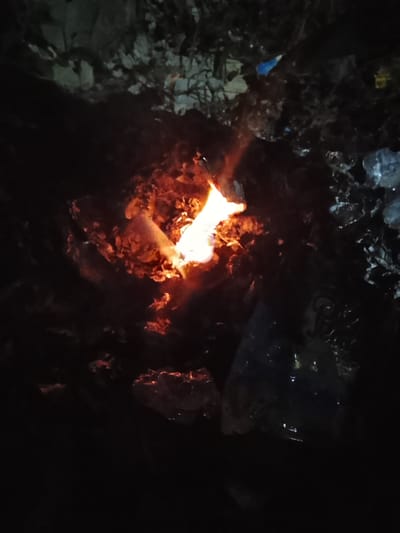 The image depicts a close-up, dimly lit view of a small fire burning amongst debris. The flames are a bright orange and yellow, casting an orange glow on the surrounding material, which appears to be a mix of dry leaves, twigs, and possibly some plastic waste. The environment is dark and appears to be outdoors, with some larger, indistinct shapes in the background that could be rocks or rubble. The overall impression is of a makeshift fire in a neglected or disused area. There is no visible indication of the specific location within Rumuokwuta, Nigeria, beyond the general context provided. There are no people or animals visible in the image.