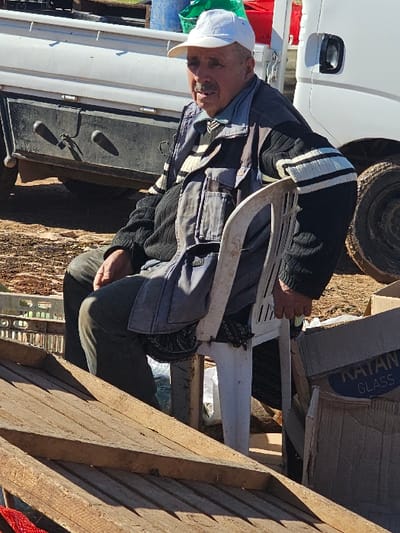 An older man with a white cap sits on a white plastic chair outdoors. He wears a layered outfit, including a blue jacket over a black sweater with white stripes. He is surrounded by clutter, including cardboard boxes, a white vehicle, and wooden planks. The lighting suggests a bright, sunny day. His expression is neutral, and he appears to be resting.