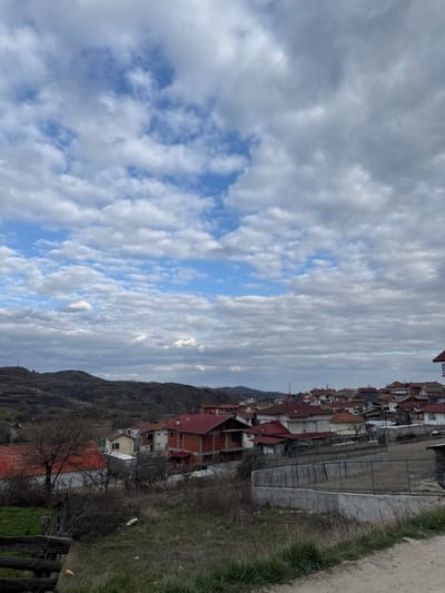 The image depicts an elevated view of Breznitsa, Bulgaria, under a partially cloudy sky displaying white and grey cumulus formations with sections of clear blue. The background consists of rolling hills sparsely covered with dry, muted green and brown vegetation. The midground features a dense residential area composed primarily of two-story houses, characterized by red-tiled roofs and light-colored or exposed brick facades, including one structure visibly under construction. Properties are separated by a mix of fences and concrete retaining walls. Scattered bare trees and dormant shrubs are visible. The immediate foreground shows a dirt path on the right, bordered by dry grass and weeds, and a segment of a wooden fence on the lower left. No individuals or active interactions are present in the scene.
