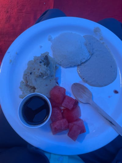 The image shows a white plate with several food items, likely a meal served in Anajpur, India. The plate contains: a round, fluffy white dish (possibly idli); a smooth, light-colored sauce; a portion of rice dish with visible spices; chunks of red watermelon; a small container with a dark liquid (likely soy sauce or a similar condiment); and a wooden spoon. The plate is resting on fabric, with blue clothing visible to the top and right, and reddish fabric on the left. The plate appears to be held or placed within a lap. There are no people distinctly visible in the image except by the clothing.
