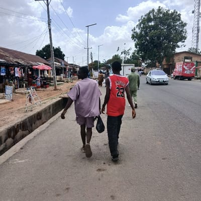 Two men are walking down a paved road in Kufang, Nigeria, located at the coordinates 9.863 latitude and 8.867 longitude. The man in the lead is wearing a red basketball jersey with the number 23 on the back. The man behind him is wearing a long-sleeved, light purple shirt. The man in the lead is carrying a dark bag over his left shoulder, while the man behind him is carrying a dark bag in his right hand. There are several businesses on the left side of the road. A small silver car is traveling towards them on the road. There are several power lines running across the top of the image. The road is flanked by a concrete curb and drainage ditch on the left, and a dirt shoulder on the right. There is vegetation growing along the sides of the road and some trees in the distance. The sky is partly cloudy.