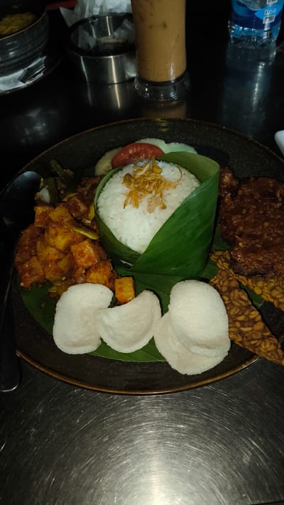 A detailed, close-up photograph captures an Indonesian meal served on a dark, textured plate, resting on a brushed metallic table surface. Central to the plate, on a green banana leaf liner, is a conical mound of white rice, partially wrapped in a fresh green banana leaf and topped with crispy fried shallots. To the left of the rice is a serving of *Sambal Goreng Kentang*, characterized by diced potatoes coated in a vibrant orange-red chili sauce, with subtle green elements interspersed. A metallic spoon handle is visible on the far left edge of the plate. To the right of the rice, several elongated pieces of fried *tempeh* are arranged alongside a substantial portion of a dark, rich-brown dish, likely a meat or poultry stew, with a thick, glossy sauce. Three white, roundish, puffy *kerupuk* (crackers) are positioned in the immediate foreground. Behind the rice, slices of fresh cucumber and red tomato are visible, serving as a garnish. In the blurred background, on the same metallic table, are a tall glass containing an iced light brown beverage, a plastic water bottle with a blue label, a dark bowl containing yellowish matter, and a metallic cylindrical container, possibly an ashtray, with discarded paper. The overall scene is dimly lit, consistent with an indoor dining environment. No individuals are present in the frame.