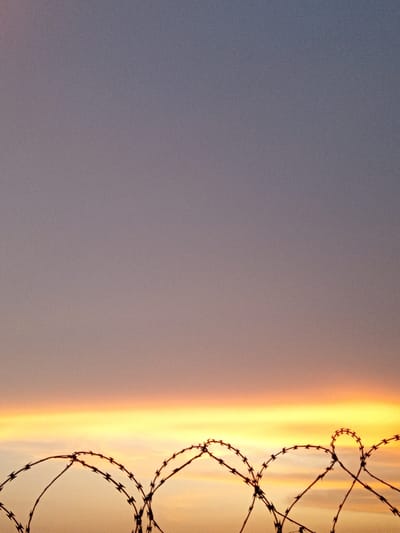 The scene in Olambe, Nigeria, depicts a sky transitioning from a muted purple-grey at the top to a vibrant horizontal band of orange and yellow light near the horizon, indicative of either sunrise or sunset. Silhouetted against this bright horizon in the foreground is a continuous run of dark, coiled concertina wire, forming multiple interlocking loops with discernible blades. No individuals, structures other than the wire, or specific landmarks are present in the image.