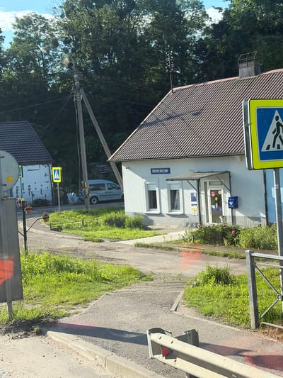 The image shows a post office in Междуречье, Russia, on a sunny day. The post office building is light-colored with a brown tiled roof and a dark blue sign above the entrance that reads "ПОЧТА РОССИИ" (Russian Post). A blue mailbox is mounted on the exterior wall near the entrance, and a transparent awning extends over the doorway. Two windows with white blinds are visible on the front of the building. To the right of the building, a yellow pedestrian crossing sign with a blue background and a white silhouette of a person walking is prominently displayed on a pole. In the foreground, a concrete sidewalk curves around a grassy area. Across the street, another building with a dark tiled roof and white walls is partially visible, as well as a lamppost with power lines running from it. A light gray van is parked in front of the white building. The environment is rural or suburban, with mature trees in the background and green grass and vegetation growing throughout the scene. The overall impression is that of a quiet, ordinary day at a local post office.