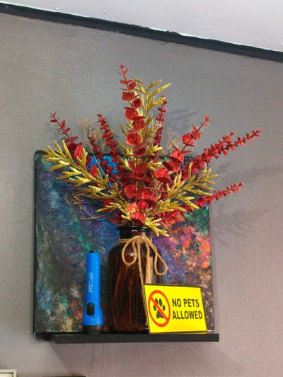 The image shows a shelf with decorative items. On the shelf, there is a brown glass vase with artificial red and gold foliage. To the left of the vase, there is a blue flashlight. Behind the vase and flashlight, there is a colorful abstract painting. In front of the painting and to the right of the vase, there is a yellow sign with a red circle and a paw print inside, indicating "NO PETS ALLOWED." The shelf is mounted on a gray wall. The overall scene appears to be indoors, likely in a home or commercial establishment. The location of Cavite City, Philippines is not visually evident from the image.