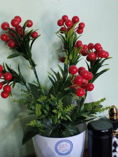The scene depicts an artificial plant with green foliage and numerous clusters of bright red, spherical berries, contained within a white, geometrically faceted pot. The pot features a circular sticker with a blue border and white background, bearing text "F.N.B TEXNOPLAST" and the Uzbek phone number +998 90 626 77 70. The plant is positioned against a light greenish-blue painted wall. To the right of the pot, a black shaker with a perforated top and a portion of another decorative item with a gold-colored cap are partially visible. No individuals are present. The environment is an indoor setting in Yangiariq, Uzbekistan.