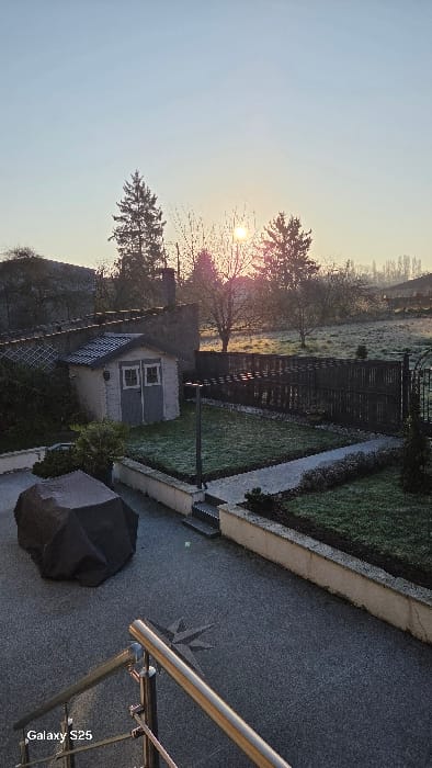 The scene depicts an outdoor residential or semi-rural garden area in Ottonville, France, captured during early morning on a clear, frosty day. The low sun, positioned brightly on the horizon behind trees in the background, casts long shadows and illuminates widespread frost across the lawns and fields.
In the immediate foreground, a metallic railing, possibly stainless steel, is visible on the lower right. Below and to the left, a dark, granular paved surface features a dark, tarp-covered object, likely outdoor furniture, and a light-colored star-shaped inlay. A small, square light-colored object with a green top is also present on the pavement.
The midground comprises a multi-level garden enclosed by low, light-colored retaining walls. Sections of green lawn are visibly covered in frost, and a light-colored paved pathway borders one area. A freestanding light-colored wooden garden shed with a dark grey gabled roof and two double doors, each featuring two small windowpanes, stands prominently on the left. A dark grey lamppost with a rectangular light fixture is centrally located on a lawn section. A dark, slatted fence forms the rear boundary of the garden, with an ornate metal gate on its right side. Various shrubs and small bushes are planted within the garden beds.
The background reveals an expansive, frost-covered field or meadow stretching towards the horizon. Scattered throughout this area are bare deciduous trees and taller evergreen coniferous trees. The sun is a bright, circular disc low on the horizon, partially obscured by tree branches, imparting a warm orange glow to the atmosphere. A stone building or wall with a dark roof is partially visible on the far left, while distant features are silhouetted against the bright sky on the right. No individuals, animals, or active interactions are present in the scene.