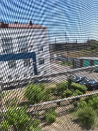 The image is a view through a screen, likely a window screen, of an urban landscape on a clear day. In the foreground, the screen mesh is clearly visible, creating a grid pattern over the scene.

The dominant feature in the midground is a white building with a red-tiled roof. This building appears to be multi-story, with visible windows and a blue accent band. To the left of this building, a portion of another structure is visible.

Behind the buildings, a parking lot or open area is present, with several dark-colored vehicles visible. The vehicles appear to be cars.

The background shows a hazy sky with a bright blue hue. In the distance, a tall industrial chimney or smokestack is visible against the sky, suggesting an industrial area. Greenery, likely trees or bushes, can be seen scattered throughout the scene, particularly in the midground and foreground. The overall impression is an outdoor scene in a built-up area, possibly on the outskirts of a city, with some industrial elements present.

There are no people, actions, or interactions depicted in the image. The scene is static and observational. The implied location of Temirtau city administration, Kazakhstan, suggests that the view might be from a building associated with the city's administration or a nearby structure looking out towards the city. However, there are no specific markers in the image to confirm this precise location.