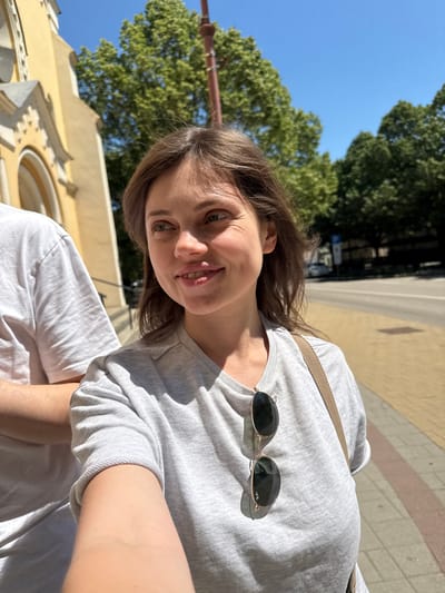 A selfie depicts a fair-skinned woman with shoulder-length light brown hair and green eyes in Siófok, Hungary. She wears a grey t-shirt with sunglasses resting on the chest area. The photo includes parts of buildings and trees indicating an outdoor setting. The image hints at possible street scenes given the background elements present.