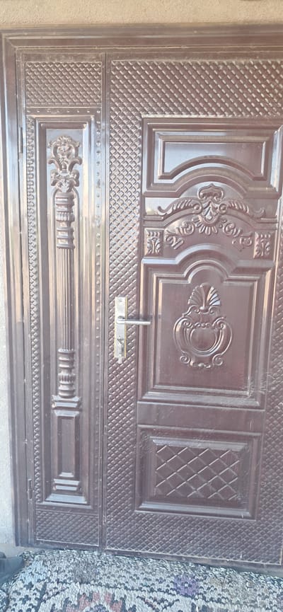 The image shows a closed double door made of dark brown metal. The doors are ornately decorated with raised patterns. The left door has a vertical decorative element resembling a fluted column with a sculpted top. The right door has several panels with intricate designs, including a shell-like motif and swirling patterns. A silver-colored door handle with a keyhole and a key is visible on the right door. The doors are set against a textured, light-colored wall. In the foreground, a patterned rug is visible on the ground, with a portion of a black shoe on the far left. The scene appears to be an entrance, likely in Yazy, Turkmenistan, given the provided context.