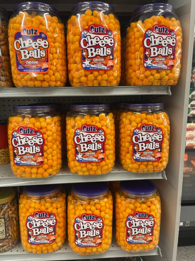 The image shows a grocery store aisle with shelves stocked with large plastic jars of Utz Cheese Balls. The jars are arranged in three rows, with three jars visible in the top and middle rows, and two jars in the bottom row. The cheese balls are orange and spherical, filling each jar. The labels on the jars are predominantly orange and white with blue accents, featuring the Utz logo and "Cheese Balls" in large lettering, along with "Cheddar" and "Real Cheese." The background reveals more shelves with other food products and a glimpse of store fixtures. The lighting suggests an indoor retail environment. The specific location in Edgartown, United States of America is unknown as the image does not provide any identifying landmarks or signs.