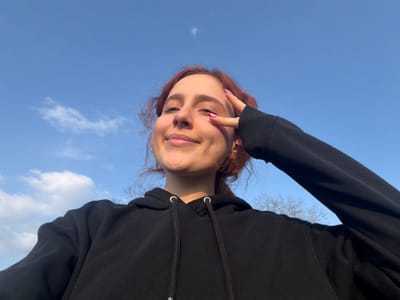 A low-angle outdoor selfie depicts a young individual, likely female, with reddish-brown hair and a smiling expression, captured in Toul, France. The subject wears a black hooded sweatshirt. Their left hand is raised to the face, forming a V-sign with pink-painted fingernails, partially obscuring the left eye. The background consists primarily of a clear blue sky with scattered white clouds. The bare branches of deciduous trees are visible in the lower right background, indicating a time of year when foliage is absent. The scene is brightly lit by natural sunlight.