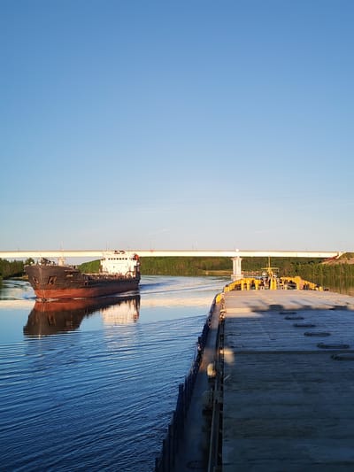 The image captures a daylight scene on a river in Ivanov Bor, Russia. In the immediate foreground, on the right side, the grey, flat deck of a large vessel, possibly a barge, extends into the frame. This deck features multiple circular access hatches. A section of a dark blue railing runs along its port side. Towards the vessel's center, a yellow structure, possibly a crane or control cabin, is visible, with indistinct figures in safety helmets on its platform.
In the midground, a dark-hulled cargo ship with a white superstructure and bridge is navigating the waterway, moving towards the viewer and slightly to the left. The name "КОПА" (KOPA) is clearly visible in white Cyrillic script on its bow. The vessel generates a distinct V-shaped wake, and its detailed reflection is present in the deep blue, calm water.
In the background, a long, light-colored bridge, supported by multiple piers, spans the river horizontally. Streetlights are affixed along the bridge deck. Both riverbanks are densely covered with green trees. A prominent bridge pier is visible on the right bank, beneath the bridge structure. Further to the right, beyond the treeline, an area of disturbed earth suggests construction or land alteration. The upper half of the image is dominated by a clear, unclouded bright blue sky, containing two thin, parallel dark lines, indicative of overhead cables, and a few minute dark specks. The scene is illuminated by direct sunlight, creating distinct shadows and bright reflections on the water.