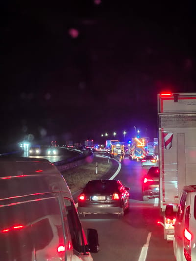The image depicts a nighttime traffic scene on a highway near Uettingen, Germany, characterized by heavy congestion and emergency vehicle presence. A significant number of vehicles are stopped on the road, indicating a traffic jam. Several emergency vehicles, including fire trucks and what appear to be police vehicles with flashing blue lights, are visible, suggesting an incident or accident has occurred. The road surface is wet, possibly due to rain, and the headlights of approaching vehicles create glare. The scene is illuminated by the headlights of the vehicles, the flashing lights of emergency services, and overhead streetlights. In the foreground, the rear of a white van or truck is prominent, with its red taillights illuminated. Several cars are also visible in the traffic jam, including a dark sedan with its license plate visible (CS 20 SBI). The overall atmosphere is one of disruption and a response to an unforeseen event on the highway.