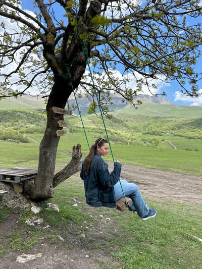 A young woman is sitting on a tree swing overlooking a mountainous landscape. The swing is attached to a tree with green rope, and there are several small wooden signs attached to the trunk. The woman is wearing jeans and a denim jacket. The background consists of green hills and a mountain range under a blue and partially cloudy sky. The location is likely Teghut, Armenia, as that is the city where this tree swing is a popular spot.